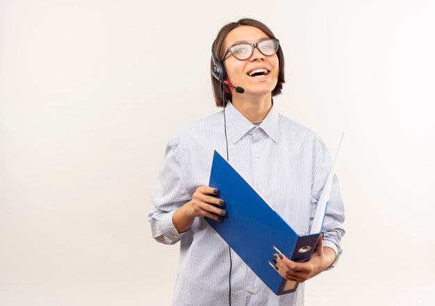 Joyful young call center girl wearing glasses and headset holding folder isolated on white