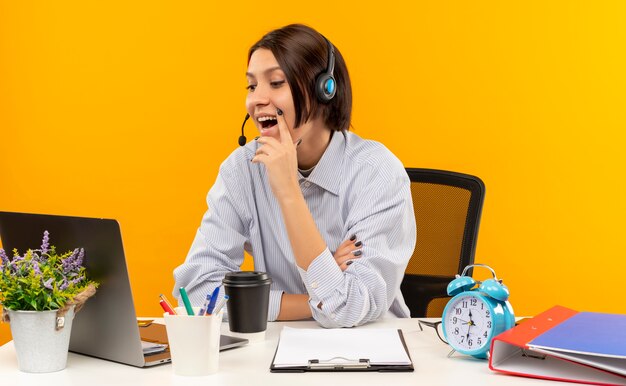 Joyful young call center girl wearing headset sitting at desk putting hand on chin looking at laptop isolated on orange