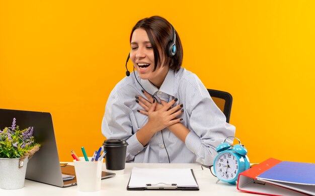 Joyful young call center girl wearing headset sitting at desk putting hands on chest with closed eyes isolated on orange