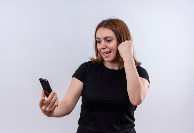 Joyful young casual woman holding mobile phone looking at it and raising fist on isolated white space