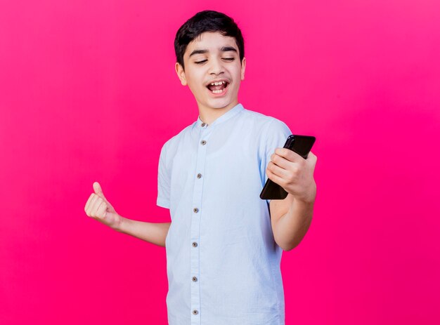 Joyful young caucasian boy holding and looking at mobile phone doing yes gesture isolated on crimson wall