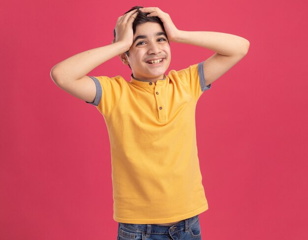 Joyful young caucasian boy  keeping hands on head isolated on pink wall