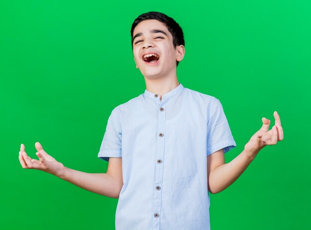 Joyful young caucasian boy looking at camera showing empty hands isolated on green background