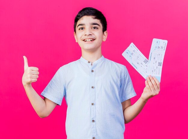 Joyful young caucasian boy looking straight holding airplane tickets showing thumb up isolated on crimson background