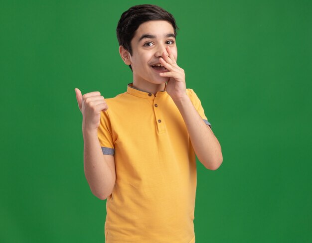 Joyful young caucasian boy putting hand on mouth pointing behind 