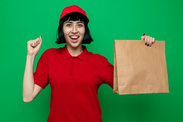 Радостная молодая кавказская доставщица держит упаковку для еды и держит кулак