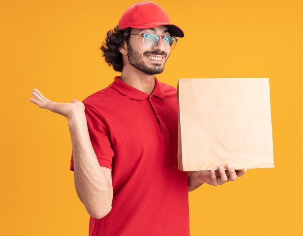 Joyful young caucasian delivery man in red uniform and cap wearing glasses holding paper package  showing empty hand isolated on orange wall