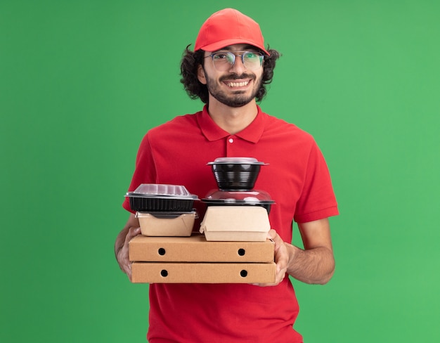 Gioioso giovane fattorino caucasico in uniforme rossa e berretto con gli occhiali che tengono pacchetti di pizza con confezioni di carta e contenitori per alimenti su di loro guardando davanti