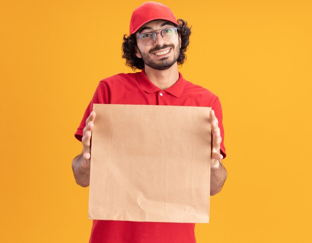 Joyful young caucasian delivery man in red uniform and cap wearing glasses stretching out paper package isolated on orange wall with copy space