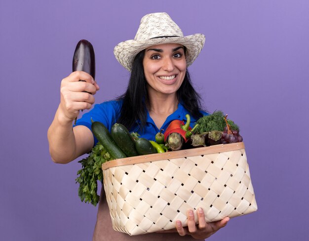 Joyful young caucasian gardener girl wearing uniform and hat holding basket of vegetables  stretching out aubergine towards camera isolated on purple wall