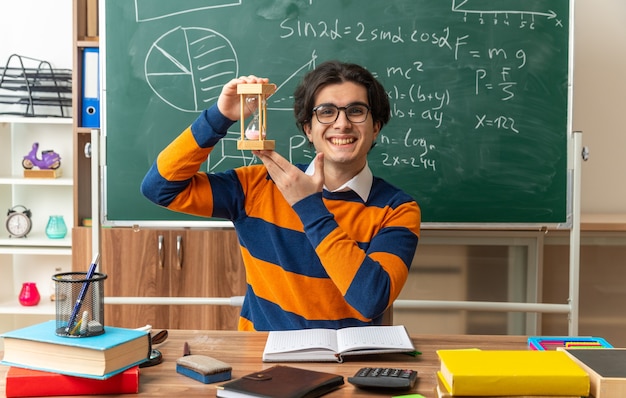 Foto gratuita gioioso giovane insegnante di geometria caucasica con gli occhiali seduto alla scrivania con strumenti scolastici in aula con clessidra