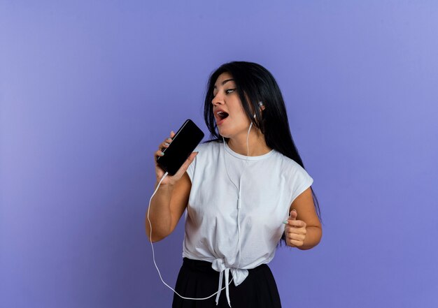 Joyful young caucasian girl on headphones holds phone pretending to sing looking at side 