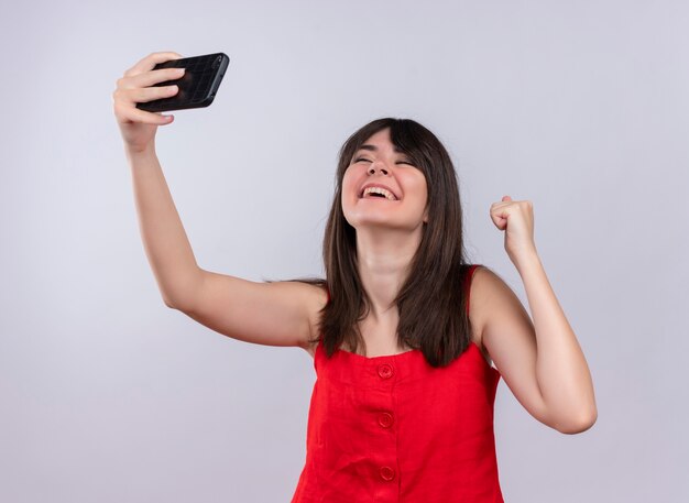 Радостная молодая кавказская девушка держит телефон и поднимает кулак, глядя вверх на изолированном белом фоне