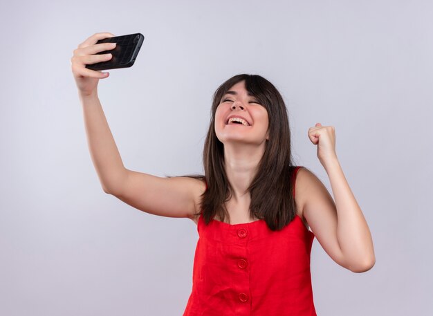 Joyful young caucasian girl holding phone and raising fist looking up on isolated white background