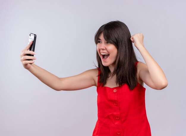 Joyful young caucasian girl holding phone and raising fist up looking at phone on isolated white background