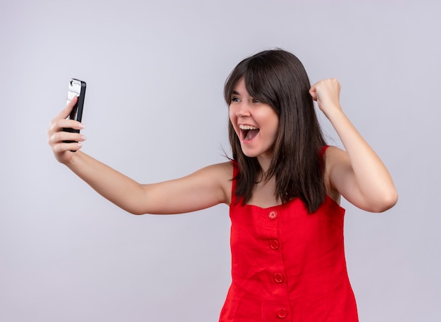 Радостная молодая кавказская девушка держит телефон и поднимает кулак, глядя на телефон на изолированном белом фоне