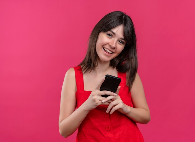 Радостная молодая кавказская девушка держит телефон обеими руками и смотрит в камеру на изолированном розовом фоне с копией пространства