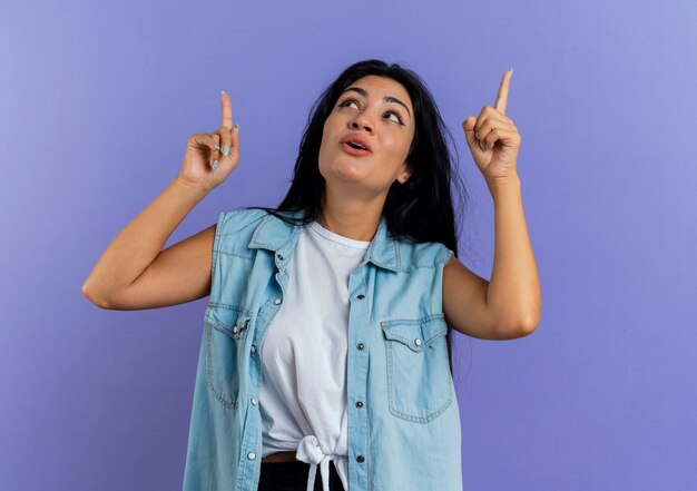 Joyful young caucasian girl looks and points up with two hands isolated on purple background with copy space