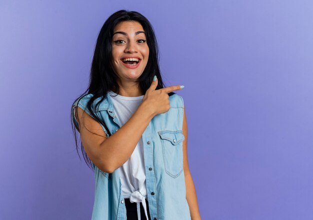 Joyful young caucasian girl points at side looking at camera isolated on purple background with copy space