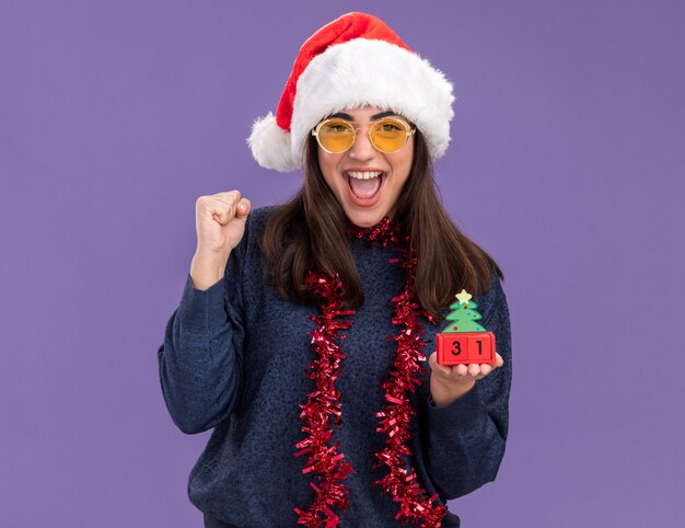 Joyful young caucasian girl in sun glasses with santa hat and garland around neck keeps fist and holds christmas tree ornament isolated on purple wall with copy space