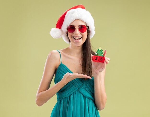 手でクリスマスツリーの飾りを保持し、指しているサンタの帽子とサングラスのうれしそうな若い白人の女の子