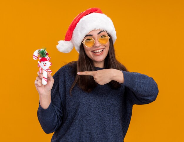 joyful young caucasian girl in sun glasses with santa hat holds and points at candy cane isolated on orange background with copy space