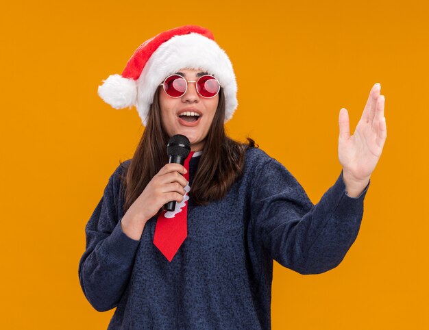 Joyful young caucasian girl in sun glasses with santa hat and santa tie holds mic and stands with raised hand isolated on orange wall with copy space