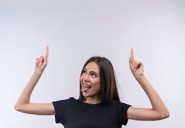 Joyful young caucasian girl wearing black t-shirt points to up showing tongue on isolated white wall