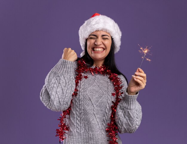 Joyful young caucasian girl wearing christmas hat and tinsel garland around neck holding holiday sparkler doing yes gesture with closed eyes isolated on purple wall