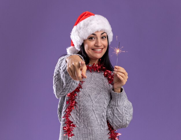Радостная молодая кавказская девушка в рождественской шапке и гирлянде из мишуры на шее держит праздничный бенгальский огонь, изолированным на фиолетовой стене с копией пространства
