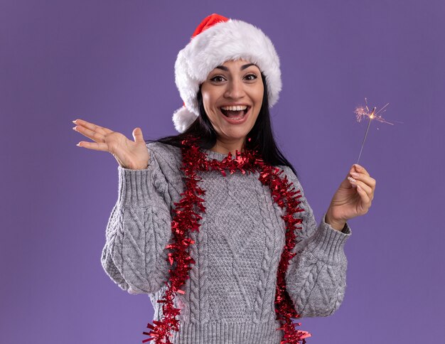 Joyful young caucasian girl wearing christmas hat and tinsel garland around neck holding holiday sparkler looking at camera showing empty hand isolated on purple background