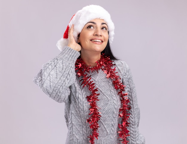 Joyful young caucasian girl wearing christmas hat and tinsel garland around neck touching hat looking up isolated on white wall with copy space