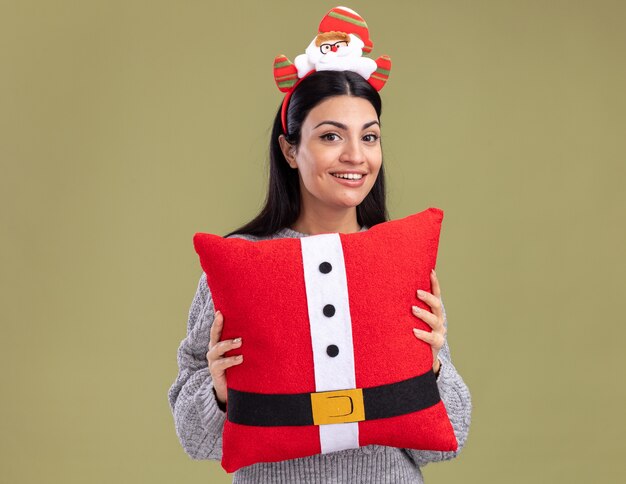 Joyful young caucasian girl wearing santa claus headband holding santa claus pillow looking at camera isolated on olive green background