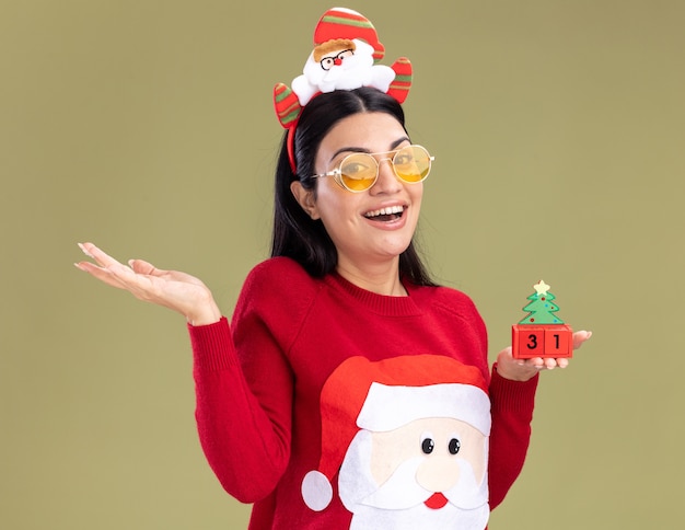 Gioiosa giovane ragazza caucasica che indossa la fascia di babbo natale e maglione con gli occhiali che tiene il giocattolo dell'albero di natale con la data che guarda l'obbiettivo che mostra la mano vuota isolata su fondo verde oliva