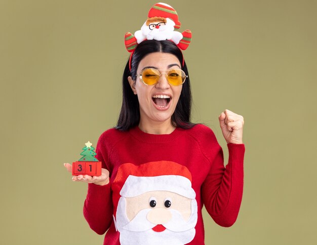 サンタクロースのヘッドバンドとセーターを着て、クリスマスツリーのおもちゃを持ったメガネをかけたうれしそうな若い白人の女の子。