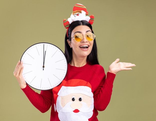 Joyful young caucasian girl wearing santa claus headband and sweater with glasses holding clock  showing empty hand isolated on olive green wall