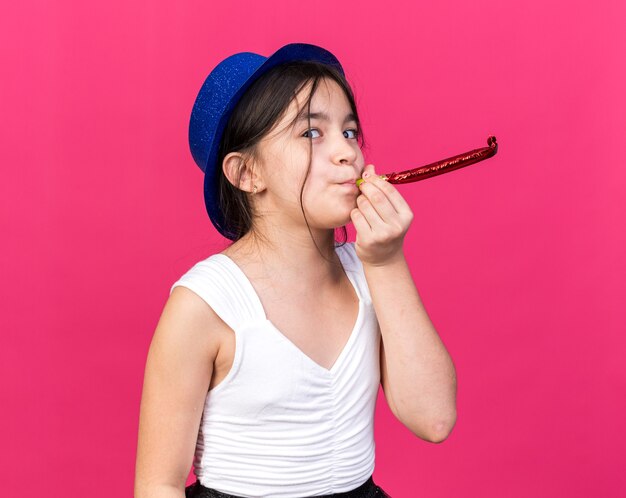 joyful young caucasian girl with blue party hat blowing whistle isolated on pink wall with copy space