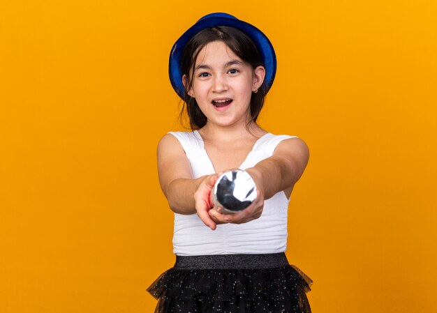 joyful young caucasian girl with blue party hat holding confetti cannon isolated on orange wall with copy space