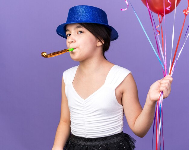 joyful young caucasian girl with blue party hat holding helium balloons and blowing party whistle isolated on purple wall with copy space