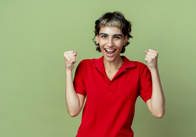 Joyful young caucasian girl with pixie haircut clenching fists isolated on olive green background with copy space