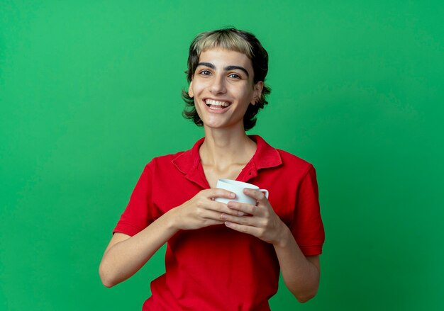 Joyful young caucasian girl with pixie haircut holding cup isolated on green background with copy space