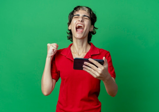 Free photo joyful young caucasian girl with pixie haircut holding mobile phone clenching fist isolated on green background with copy space