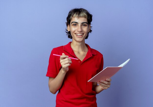 Joyful young caucasian girl with pixie haircut holding pen and note pad isolated on purple background with copy space