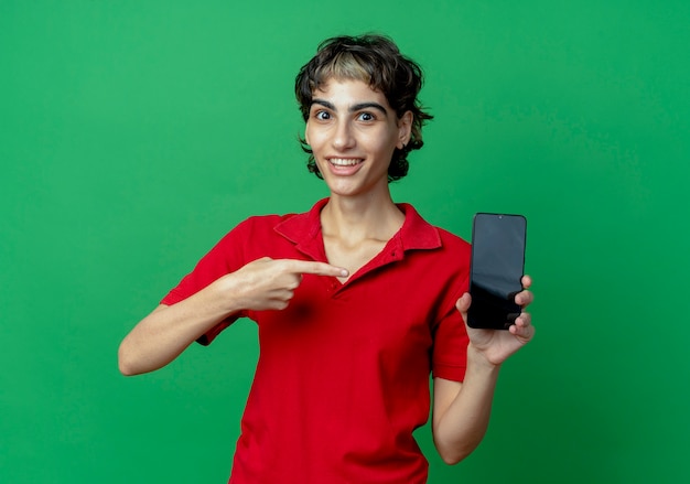 Joyful young caucasian girl with pixie haircut holding and pointing at mobile phone isolated on green background
