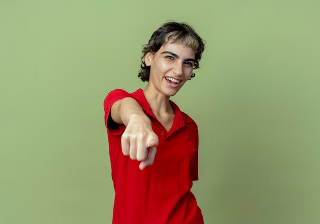 Joyful young caucasian girl with pixie haircut pointing at camera isolated on olive green background with copy space