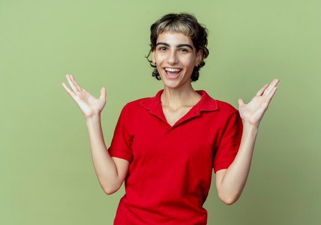 Joyful young caucasian girl with pixie haircut showing empty hands isolated on olive green background