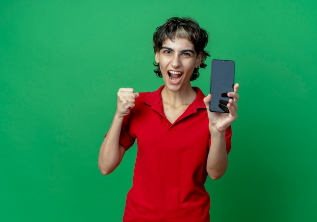 Радостная молодая кавказская девушка со стрижкой пикси показывает мобильный телефон и сжимает кулак, изолированную на зеленом фоне с копией пространства