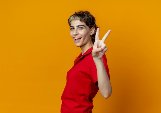 Joyful young caucasian girl with pixie haircut standing in profile view doing peace sign isolated on orange background with copy space