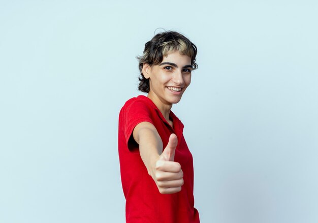 Joyful young caucasian girl with pixie haircut stretching out hand and showing thumb up at camera isolated on white background with copy space