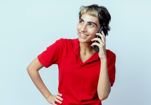 Joyful young caucasian girl with pixie haircut talking on phone looking up putting hand on waist isolated on white background